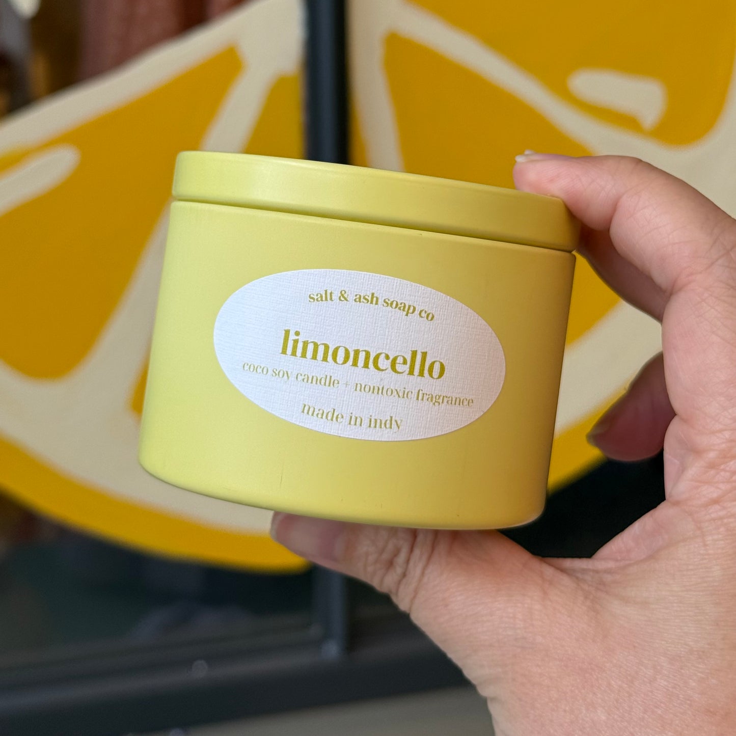 A hand holding a yellow cylindrical candle tin labeled 'limoncello' by 'salt & ash soap co', with a blurred yellow lemon graphic in the background.