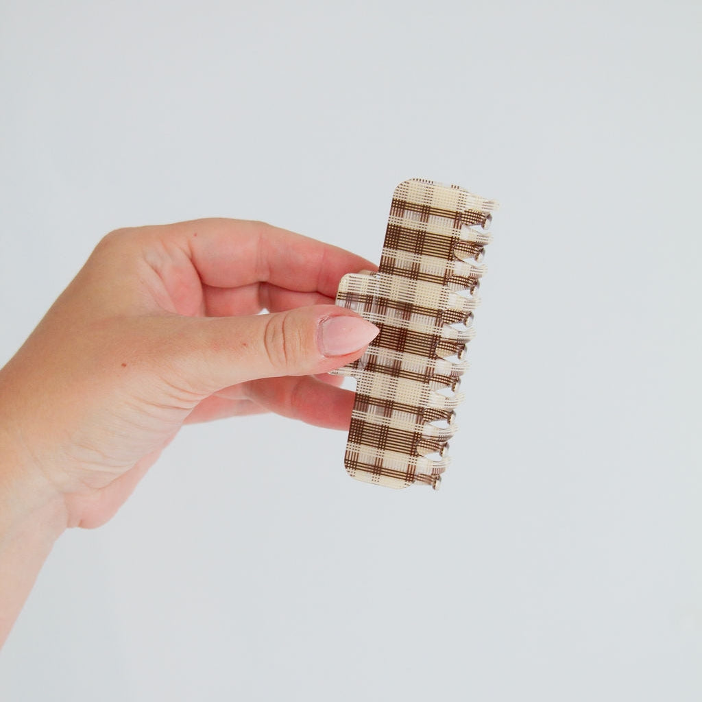 A hand holding a small hair claw clip with a brown and beige plaid pattern against a plain light gray background.