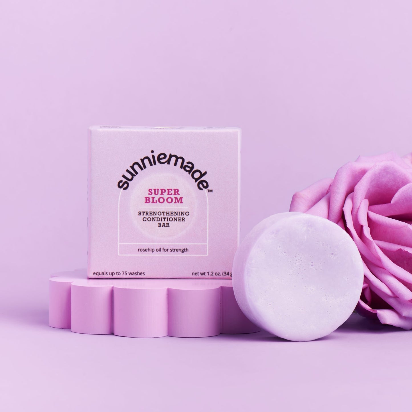 Sunniemade Super Bloom Hydrating Cleansing Oil in a pink box, shown next to a round white bar of the same oil and a pink rose on a solid lavender background.