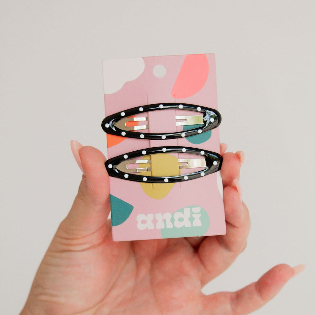 A hand holds a pink card displaying two black hair claw clips with white polka dots and pastel-colored elastic sections, against a plain light background.