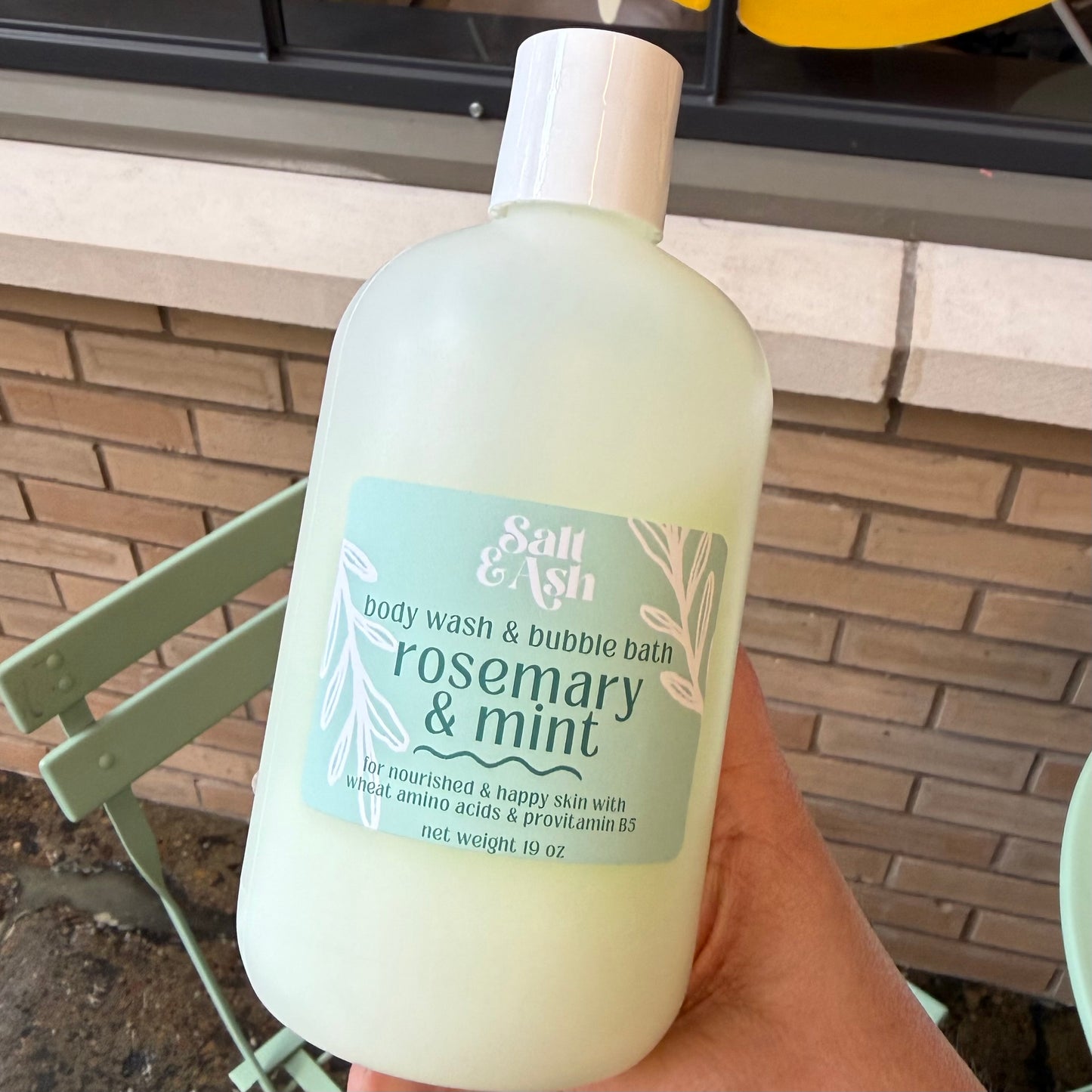 A hand holds a large frosted bottle of Salt & Ash Rosemary & Mint body wash and bubble bath, set against an outdoor patio background with a brick wall and a green chair.