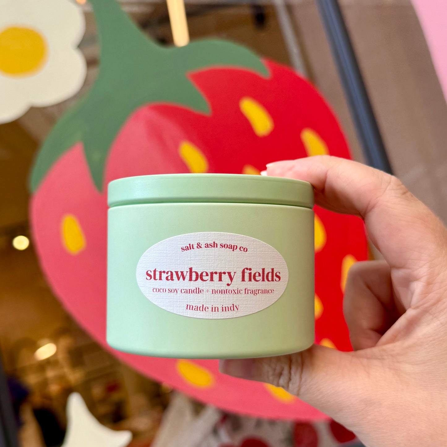 A hand holding a mint green candle tin labeled 'strawberry fields' by 'salt & ash soap co', with a large strawberry decoration blurred in the background.