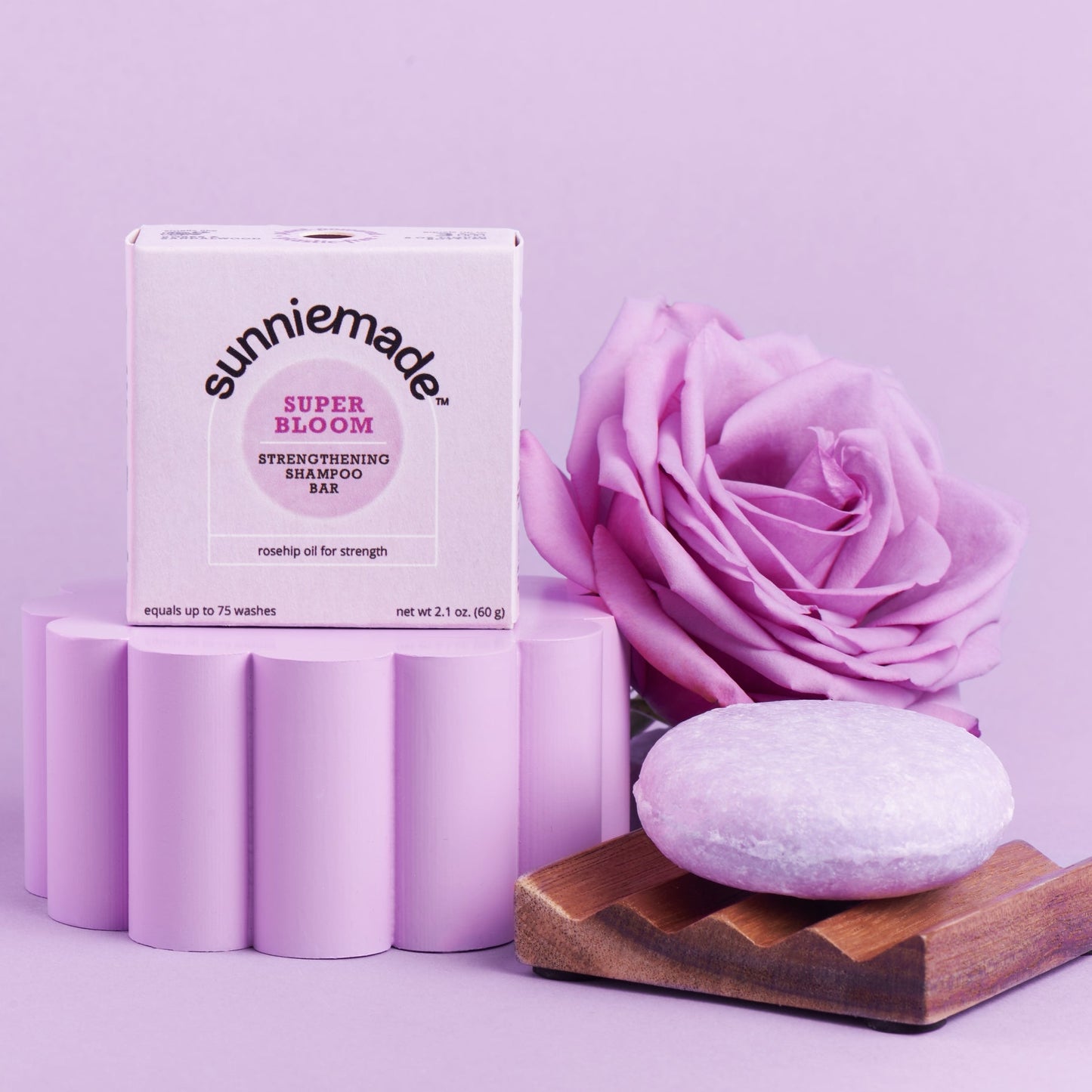 Sunniemade Super Bloom Strengthening Bar Soak set, featuring a white product box, five lavender cylindrical bars stacked on a pedestal, a large lavender rose, and a round bar of soap on a wooden soap dish, all set against a solid lavender background.