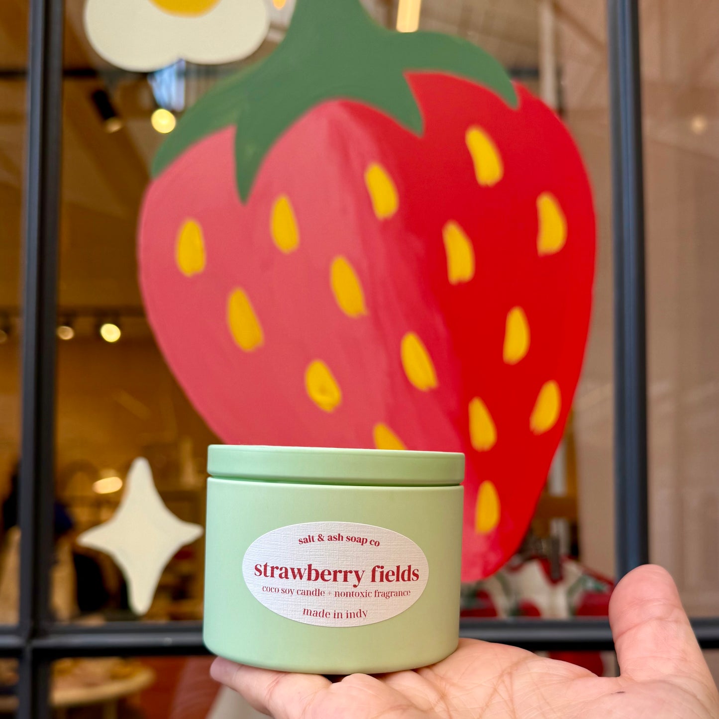 A hand holds a light green jar candle labeled 'strawberry fields' in front of a large decorative strawberry sign on a shop window.