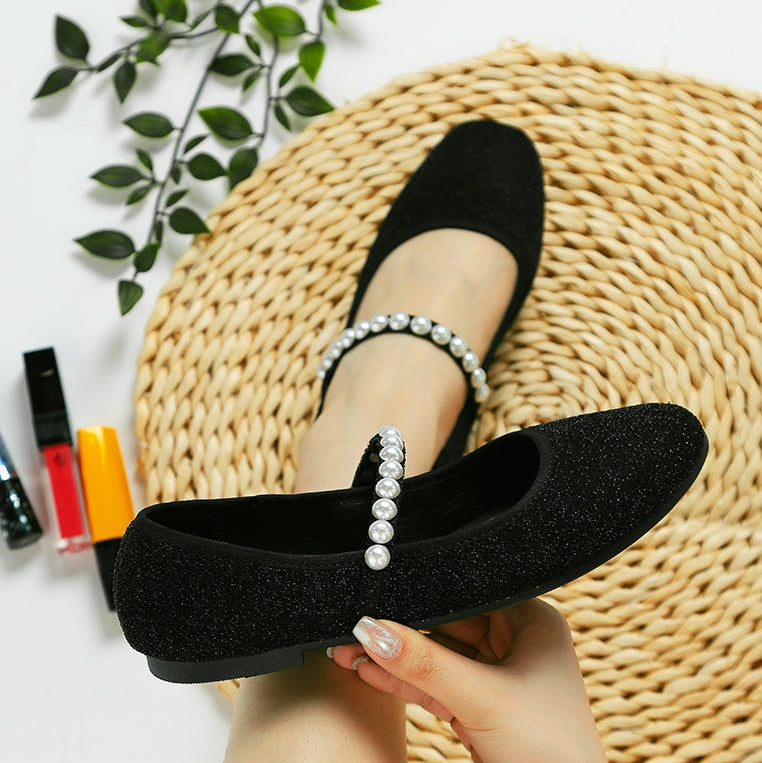 Black shoes with pearl details held by a hand on a woven mat with greenery and makeup products.