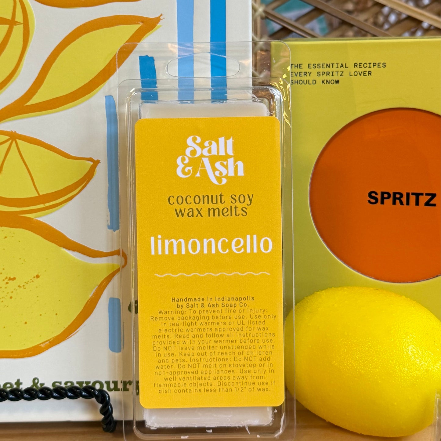 A clear plastic clamshell package of Salt & Ash coconut soy wax melts in the limoncello scent, displayed on a wooden shelf next to a yellow and orange book and a yellow lemon.