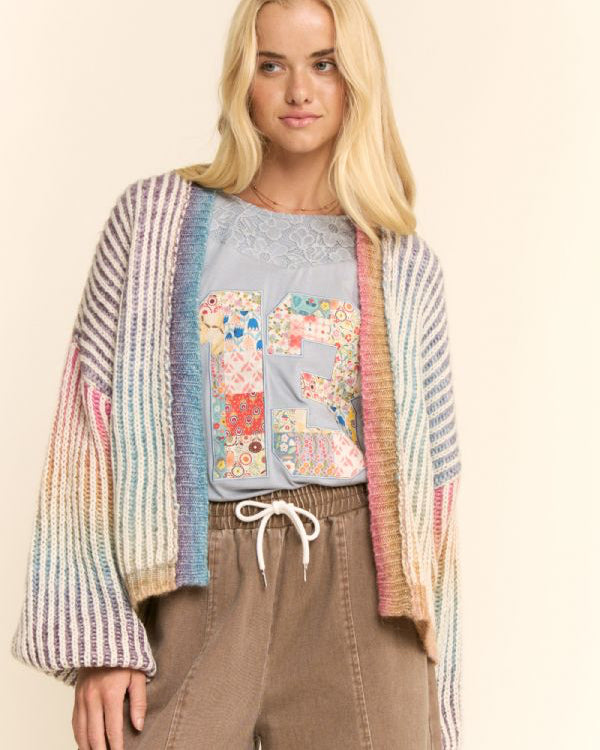 A blonde model wearing a pastel multicolor striped knit cardigan over a light blue graphic t-shirt and brown drawstring pants, set against a plain beige background.