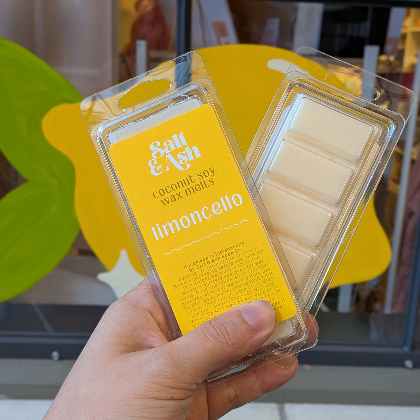A hand holds two packages of Salt & Ash coconut soy wax melts, one labeled 'limoncello' with a bright yellow label and the other showing the cream-colored wax melts inside a clear plastic clamshell pack, against a blurred storefront background.