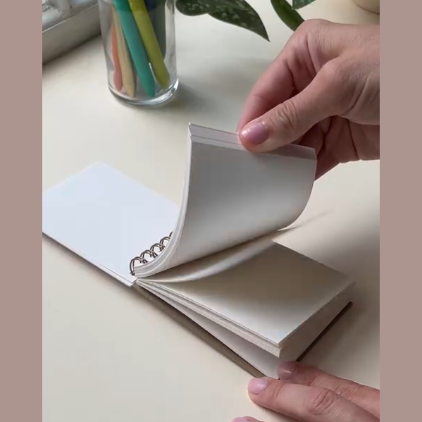 Hand holding a small open notebook on a light surface with a glass container of colorful pens in the background.