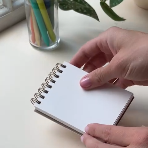 Person holding a small spiral-bound notebook on a light surface with colorful markers in the background.